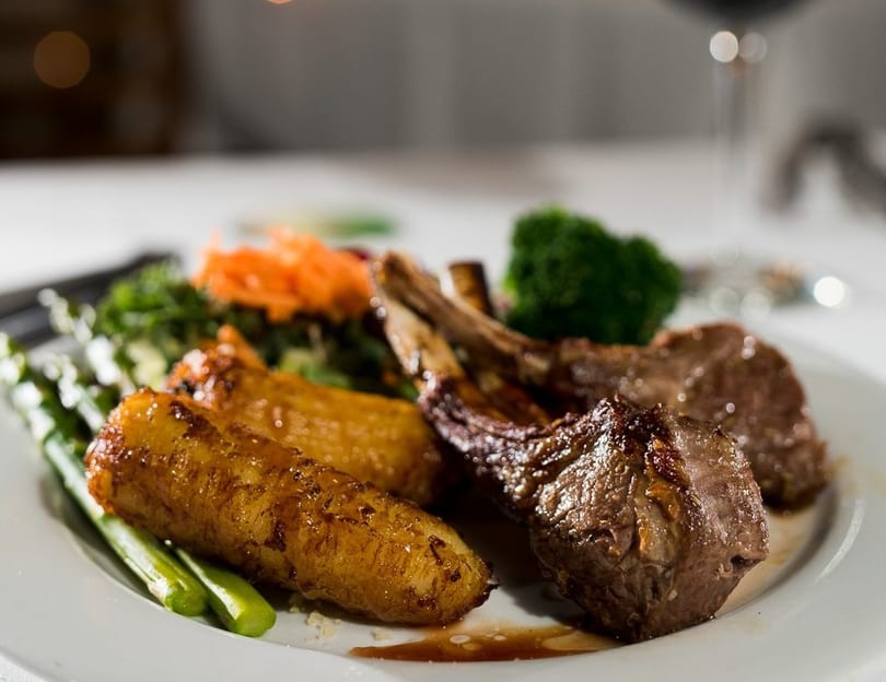 Grilled lamb chops with roasted plantains, asparagus, and broccoli on a white plate.