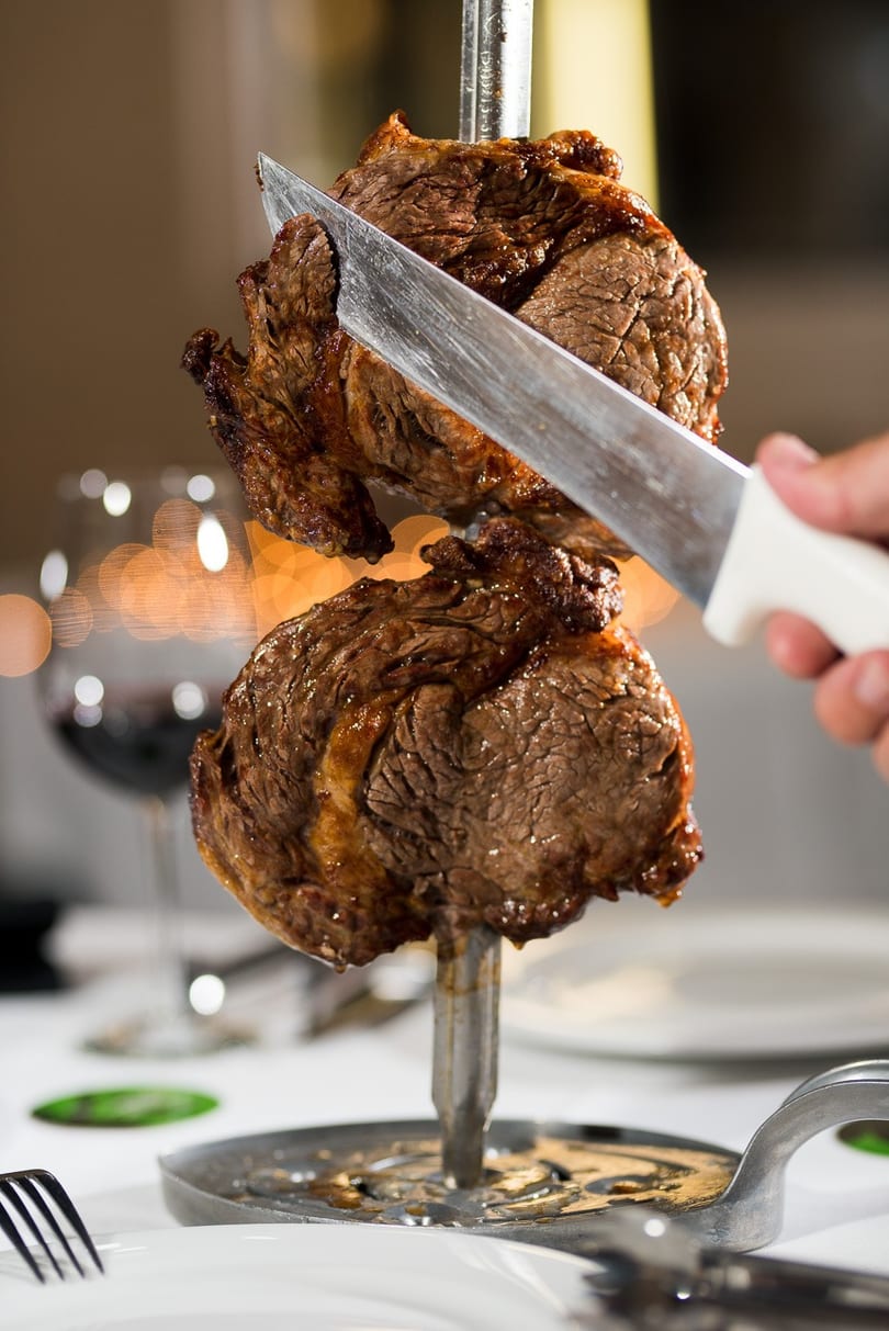 A hand slices succulent grilled meat from a skewer at a restaurant table with wine glasses.