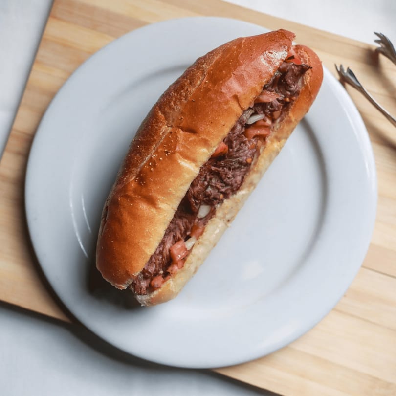 A beef sandwich with tomatoes and onions on a white plate, with tongs on the side.