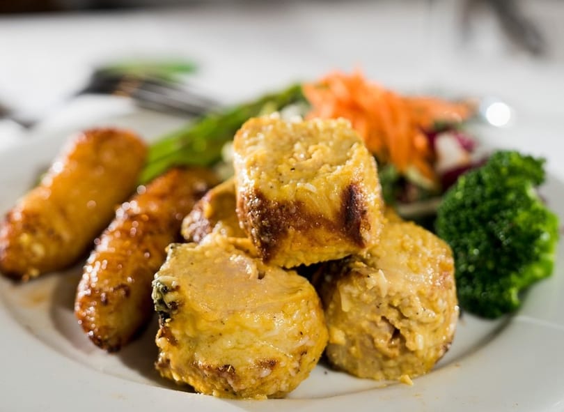 Close-up of golden-brown chicken rolls, plantains, broccoli, and salad on a white plate.