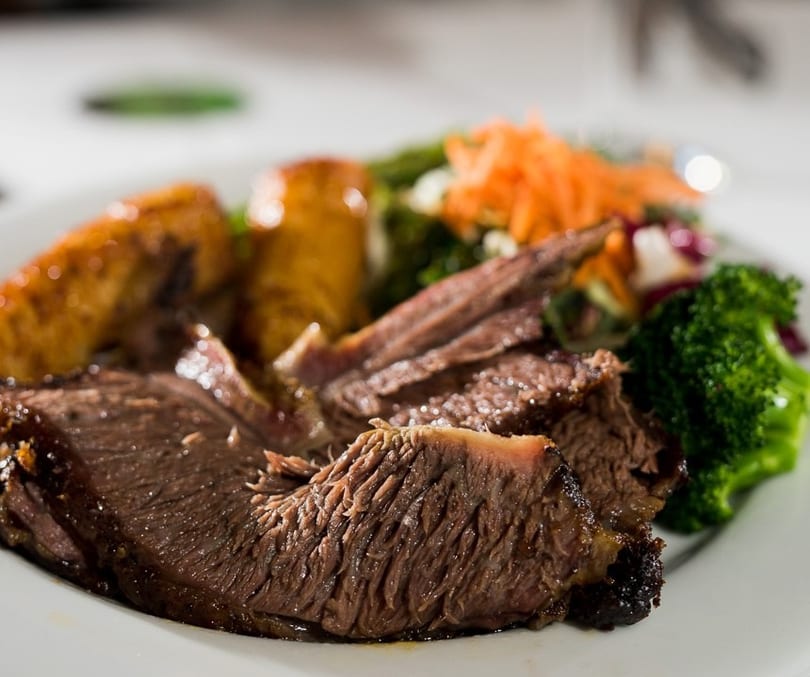 Close-up of sliced roasted beef with broccoli, shredded carrots, and plantains on a white plate.