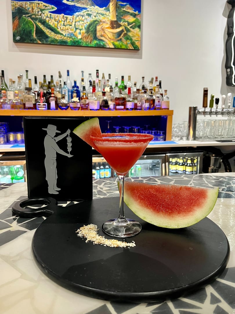 Watermelon martini with a watermelon slice and coconut flakes on a black tray at a bar.