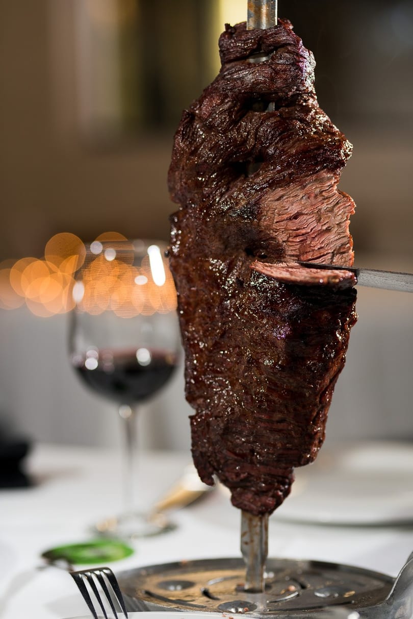A knife slices a piece of picanha from a large skewer, with a glass of red wine in the background.