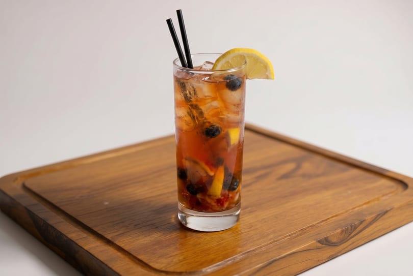 Iced tea with blueberries, lemon, and two black straws on a wooden board.