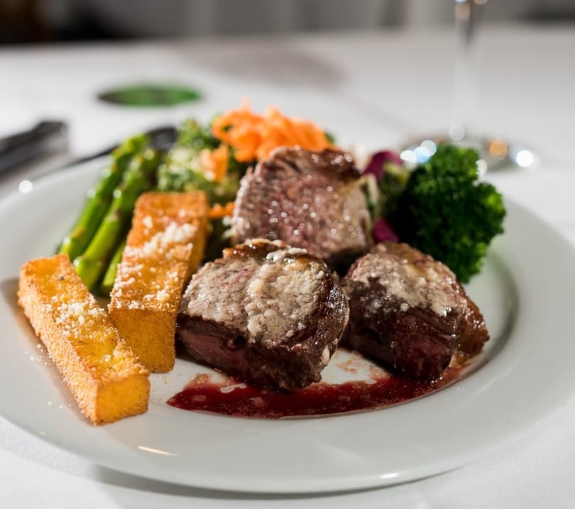 Plated steak medallions with polenta fries, asparagus, broccoli, and shredded carrots on a white tablecloth.